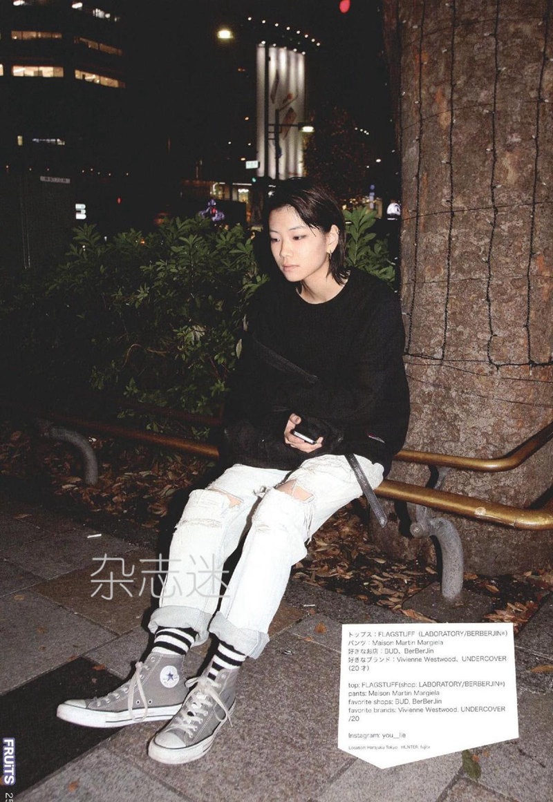 a young woman with a short haircut is captured in a street style photograph. she is sitting on a bench illuminated by streetlights at night. her outfit features a black sweater, a white tshirt, ripped white jeans, striped socks, and grey converse sneakers. text in japanese and english describing her outfit and the magazine's name "fruits" are overlaid on the image. the overall aesthetic is casual and edgy, typical of japanese street fashion.