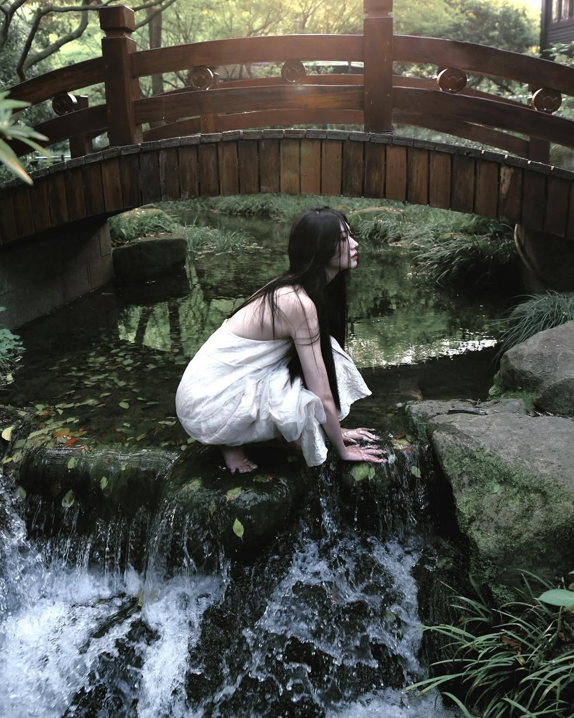 a woman with long black hair, dressed in a white dress, is sitting on mossy rocks near a waterfall. she's positioned under a wooden bridge in a lush, natural setting. the atmosphere is serene and somewhat ethereal.