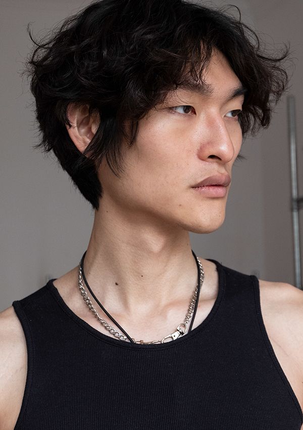 a closeup side profile portrait of a young asian male model. he has short, curly black hair, and his expression is neutral and calm. he is wearing a simple black tank top and two silver necklaces. the background is a muted gray. the overall mood is serious and fashionable.