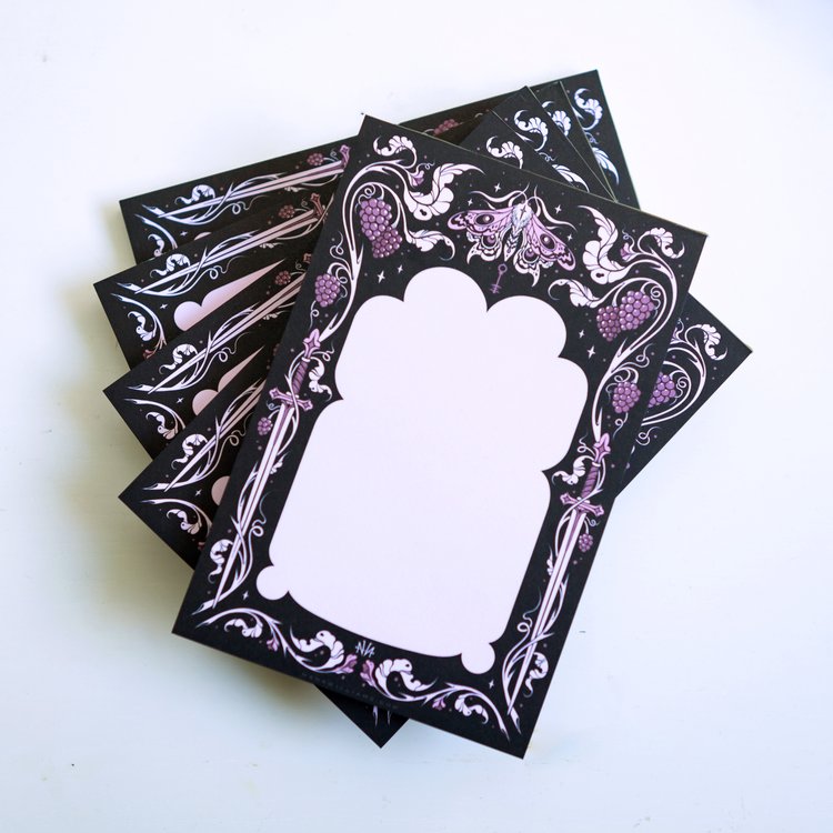a stack of black cards with decorative purple and white designs is shown. the cards feature a blank white space in the center, surrounded by a black frame with illustrations of grapes, swords, and a moth. the designs are ornate and whimsical.