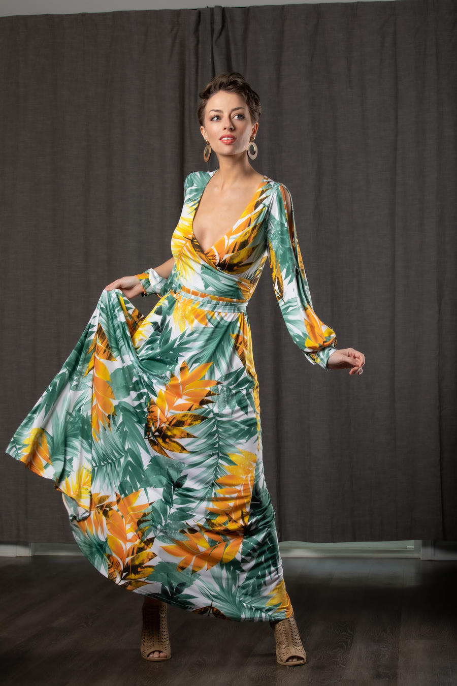 a fulllength shot of a woman in a long, floral maxi dress. the dress is primarily white with a tropical leaf print in shades of yellow, orange, and green. she is wearing sandals and has her hair styled in a short, slightly messy updo. she is standing against a dark grey curtain backdrop and is looking off to the side with a confident expression. the overall mood of the image is elegant and glamorous.