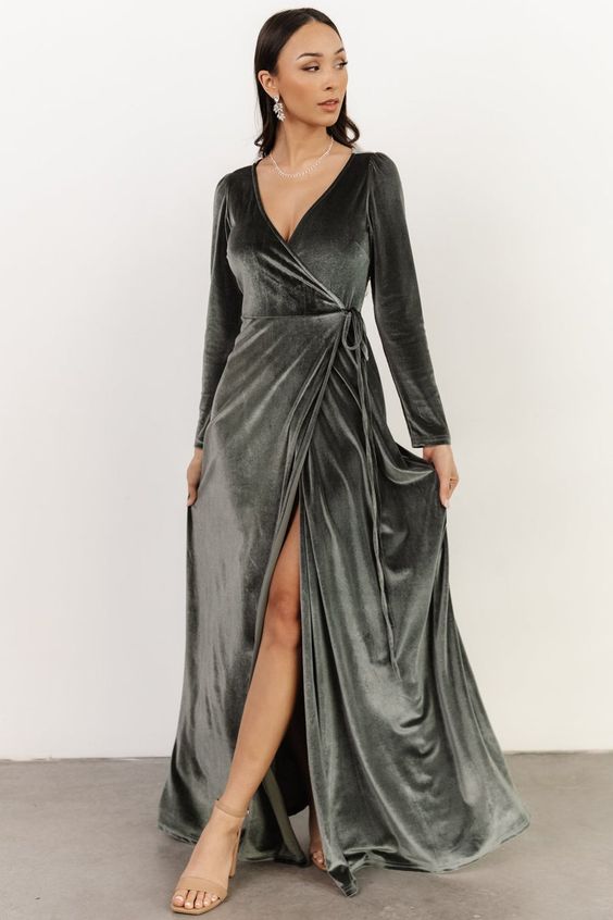 a woman is standing in a studio, wearing a longsleeve velvet wrap dress. the dress is a dark graygreen color and has a deep vneckline. the dress also has a thighhigh slit. the woman is wearing strappy heels and has her hair down. the background is white.