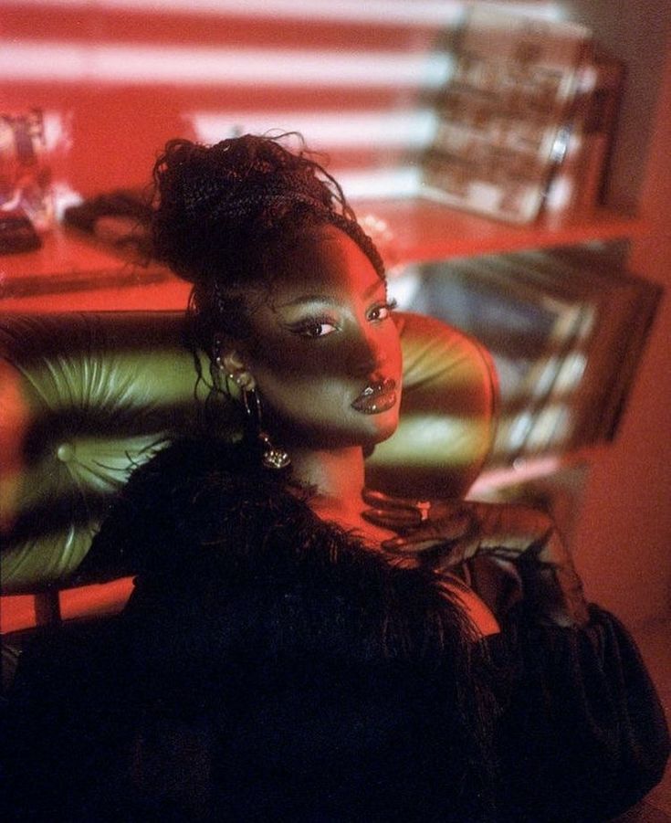 the image is a closeup portrait of a glamorous black woman with dark skin and intricately braided hair.  she's wearing a dark, possibly black, outfit with a feathery or furlike detail around the shoulders.  she has on noticeable earrings and dark gloves.  the setting appears to be an interior, possibly a dimly lit room or bar, with a reddish, almost bloodred, light casting dramatic shadows across her face and the leather chair she is sitting in. the overall mood is moody, sensual, and evocative of a film noir aesthetic, with a retro or vintage feel. the lighting and styling create a striking and powerful visual effect.