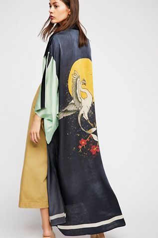 a woman is showcasing a long, elegant kimonostyle cardigan. the garment features a striking darkcolored base with a painted design of a crane in flight amidst a starry backdrop and red floral accents. the sleeves are a contrasting lighter color, adding a unique design element. the model wears the cardigan over a mustardyellow culottes and appears poised and stylish. the overall aesthetic leans towards bohemian chic, ideal for casual or more formal outings.