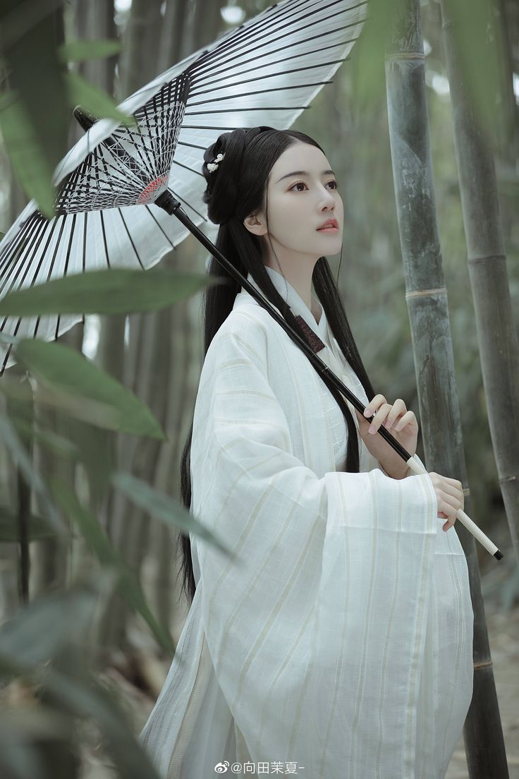 a woman in traditional chinese clothing (hanfu) stands in a bamboo forest holding a white parasol. she is wearing a long, white robe with delicate embroidery and has her long black hair pulled back in a traditional style. the forest is lush and green, and the woman is bathed in the dappled sunlight filtering through the leaves. the image is serene and peaceful, and the woman looks elegant and graceful.
