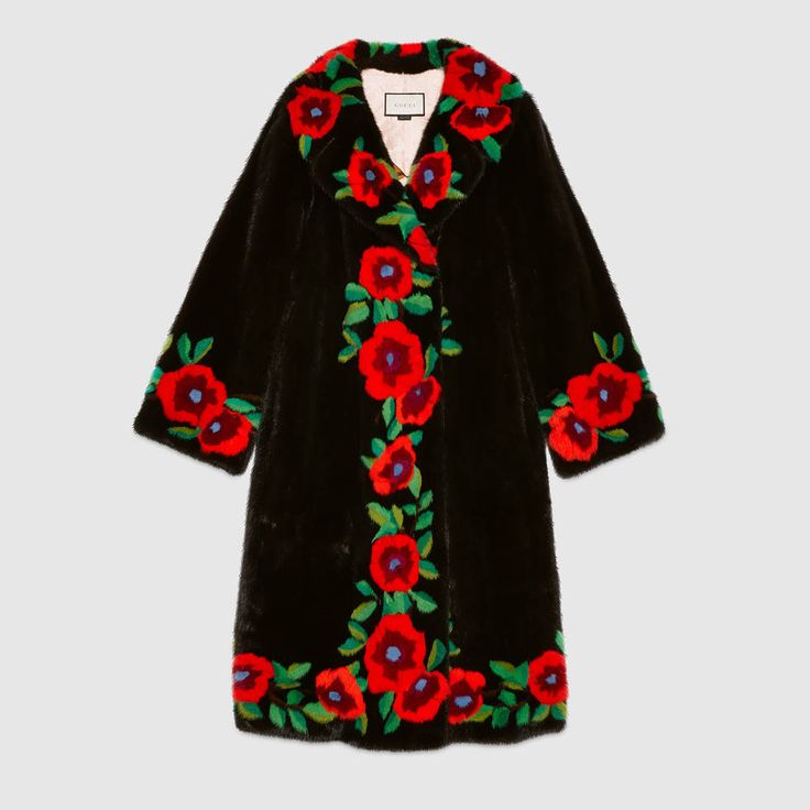 a black coat with floral embroidery is shown in a full shot. the coat is long and has a soft, furlike texture. the embroidery consists of red flowers with green leaves and blue centers, arranged along the collar, down the front, and on the cuffs. the coat appears to be a luxury fashion item, and the brand is recognizable as gucci.