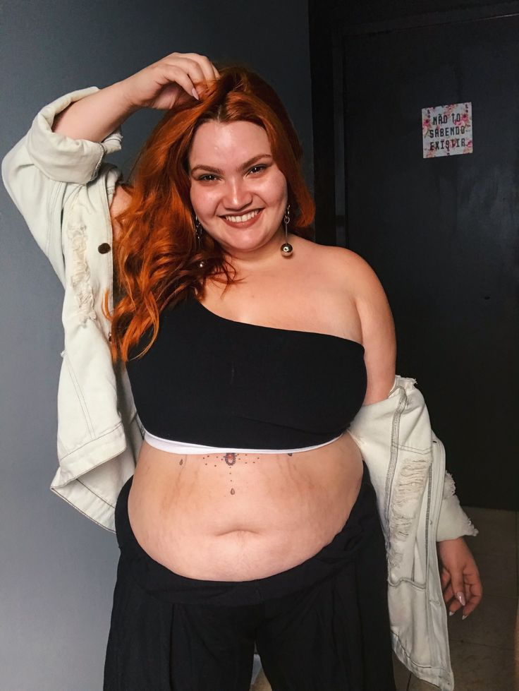 a smiling plus size woman with red hair is posing indoors. she is wearing a black one shoulder top, a white denim jacket draped over her shoulders, and dark pants. she has earrings and a tattoo on her stomach. she appears confident and happy.