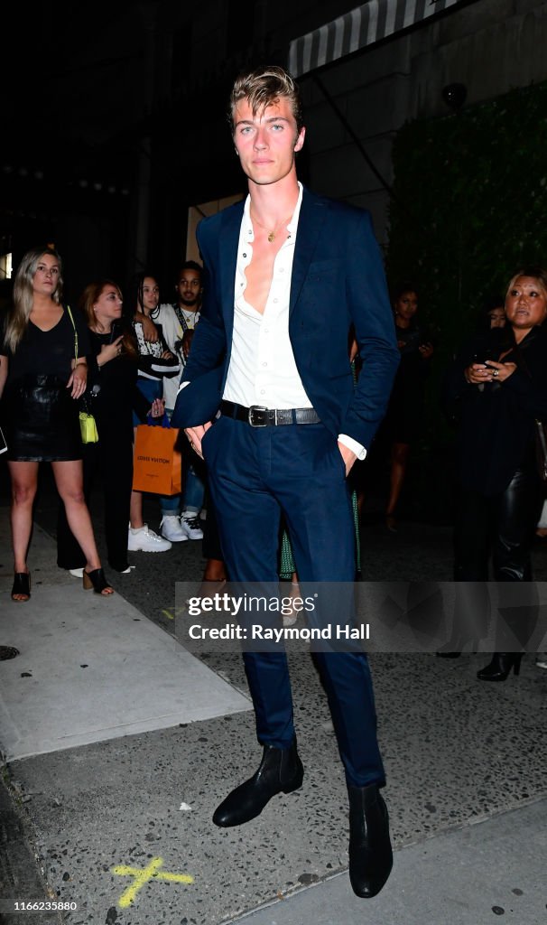 a full outdoor shot of a tall blond man in a navy suit with a white shirt, dark belt, and black chelsea boots is surrounded by other people including women and one man. the image appears to be on the street in front of a brick wall. a yellow cross is painted on the sidewalk.