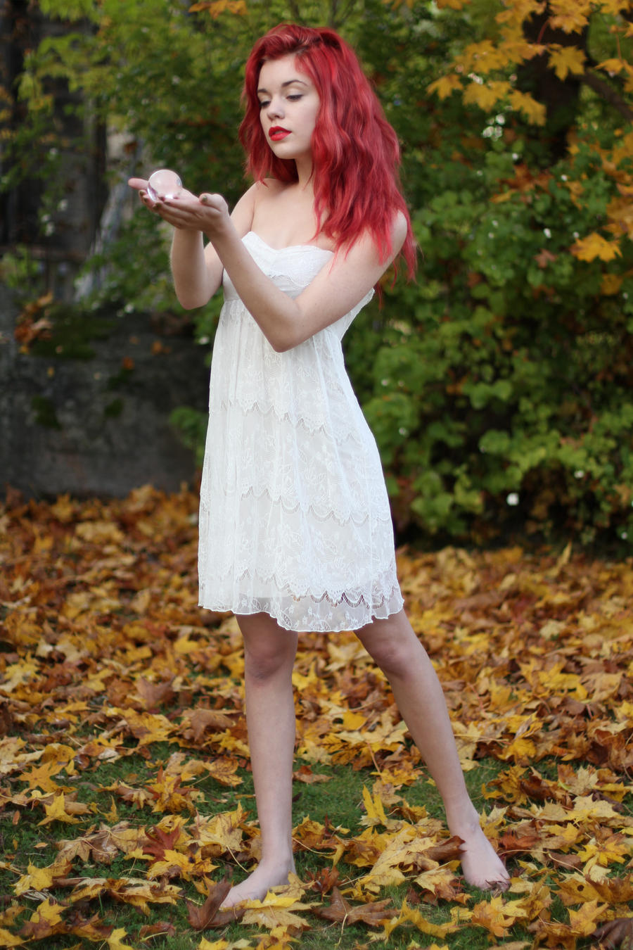 a young woman with vibrant red hair, wearing a white lace dress, stands barefoot in a park or garden covered with fallen autumn leaves. she is holding a small, lightcolored object in her hands, and her gaze is directed downward, creating a serene and peaceful mood. the image is welllit, showcasing the colors of the leaves and the woman's dress.  it appears to be a posed, professional photograph.