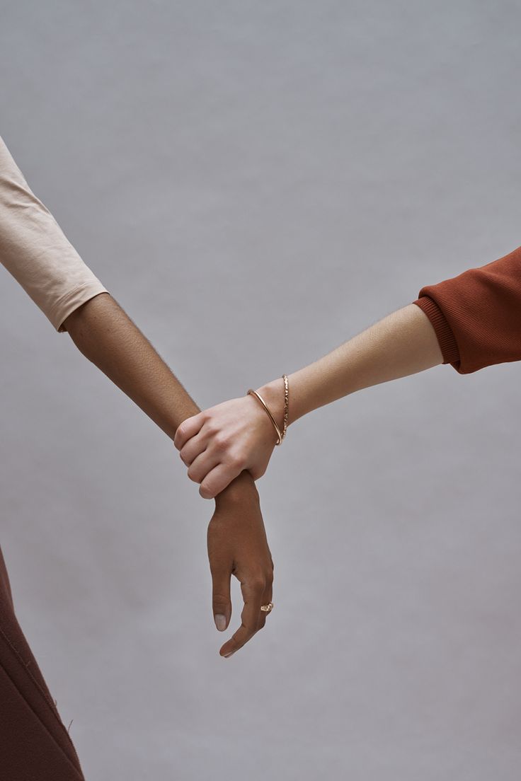 a photo of two people holding hands. the hand on the left is a brown skin person and the hand on the right is a white skin person. they're wearing bracelets and rings. the background is a light gray. the photo conveys unity and diversity.
