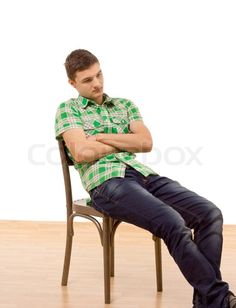 a young man is sitting in a wooden chair with his arms crossed. he is wearing a plaid shirt and jeans. he is looking down and appears to be thoughtful or sad. he is sitting in front of a white background.