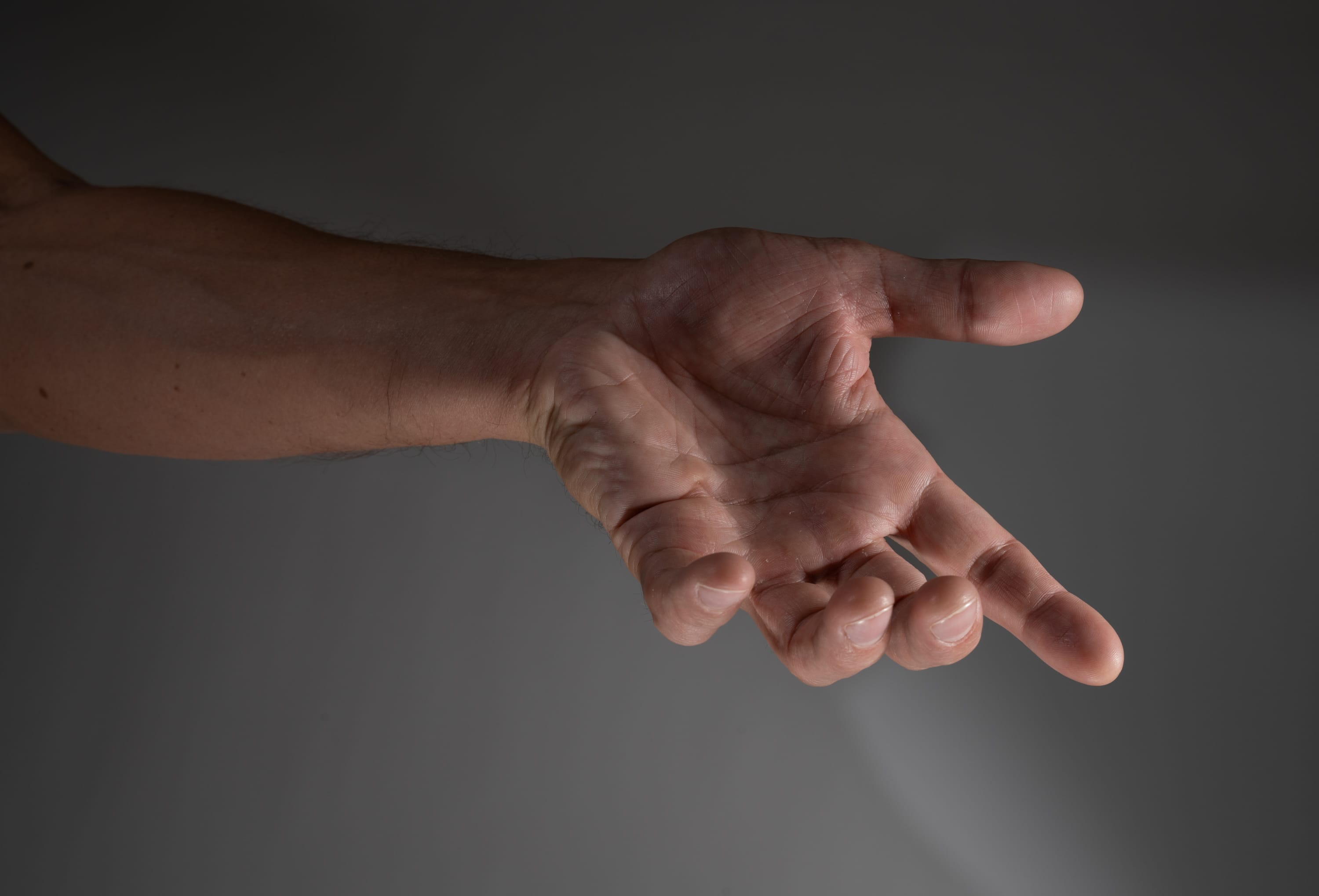 a closeup side view of a man's outstretched hand against a neutral gray background, illuminated by soft studio lighting. the hand appears to be reaching out, creating a sense of offering or asking.