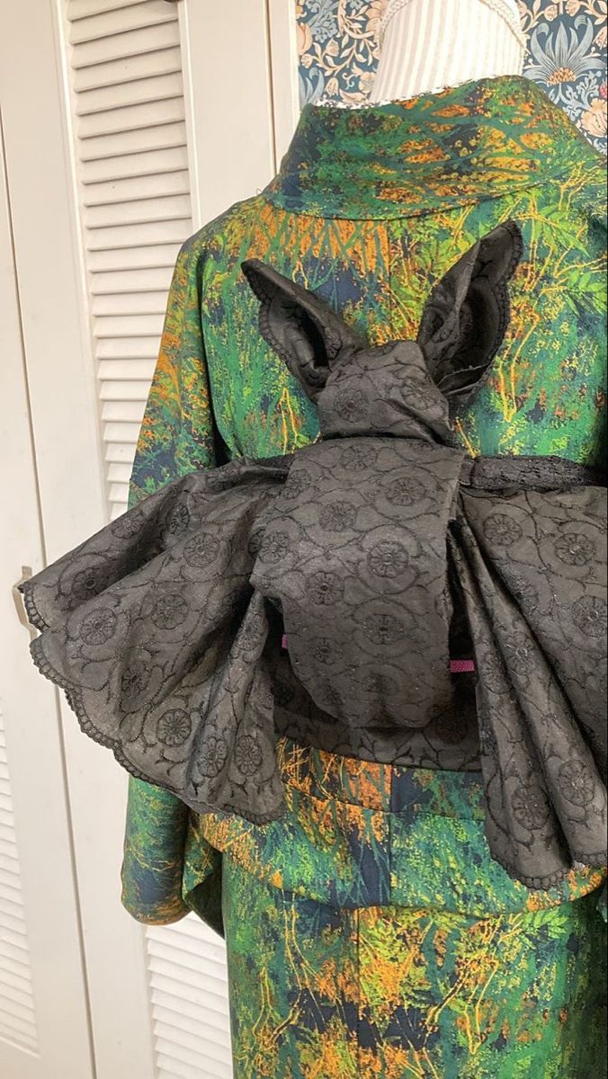 a closeup shot shows a green and gold patterned kimono with a large, black bat obi wrapped around the waist. the bat obi is made of black fabric with a subtle floral pattern. the bat's head and ears are clearly visible, while the wings are spread out on either side. the kimono has a textured, organic pattern that resembles a forest or jungle. the overall aesthetic is gothic and halloweenthemed.