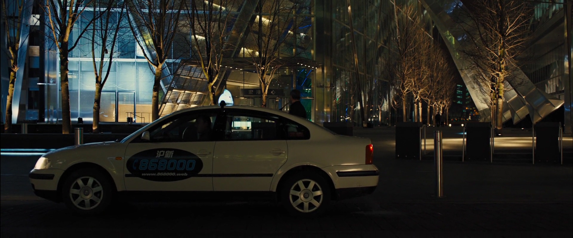 a white taxi is parked on a city street at night. the street is lined with trees and modern buildings. the scene is lit by street lights, creating a warm glow on the taxi.  a person can be seen walking on the sidewalk in the background.