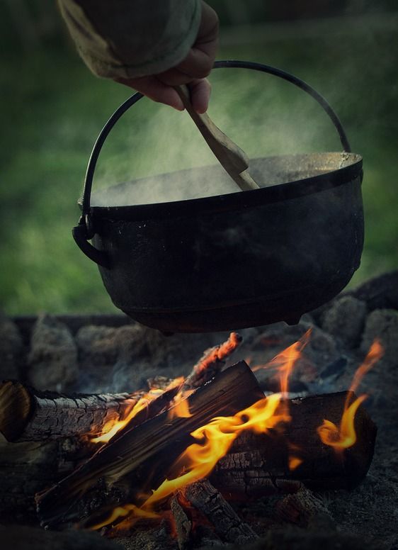 a hand stirs a cauldron with a wooden spoon over a campfire. smoke rises from the boiling cauldron and the flames licking at the firewood below. the scene takes place outdoors, with a backdrop of blurred green foliage. the image evokes a sense of medieval or rustic cooking.