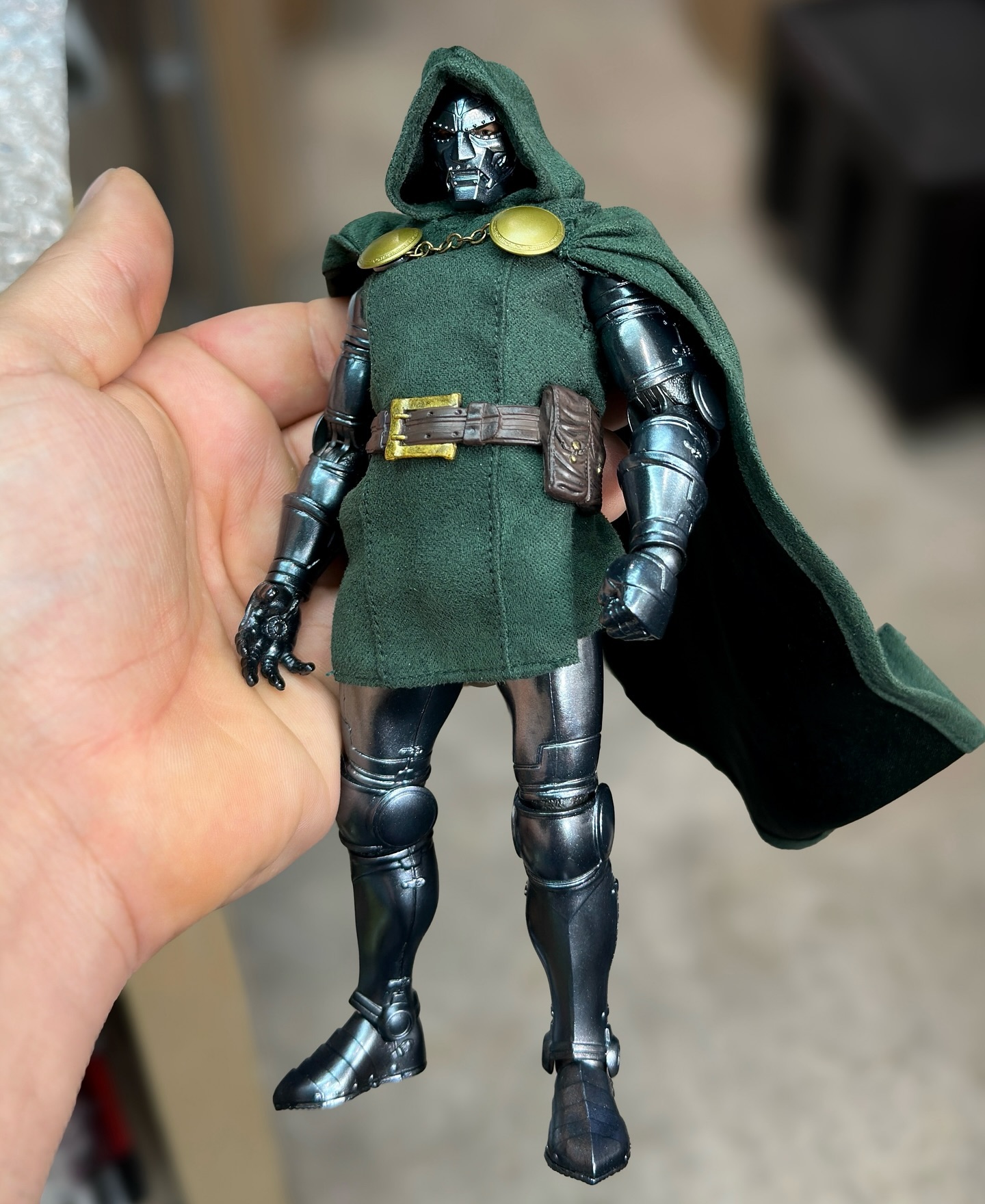 a doctor doom action figure is held in a person's hand. the figure is wearing a green cloak with a hood and metal armor. the figure's face is metallic and masked.
