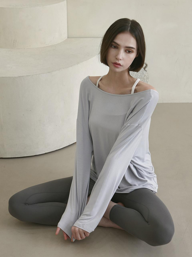 a woman is sitting in a studio shot, wearing a gray long sleeve shirt and gray leggings. she has brunette hair and short hair. the shirt is off the shoulder.
