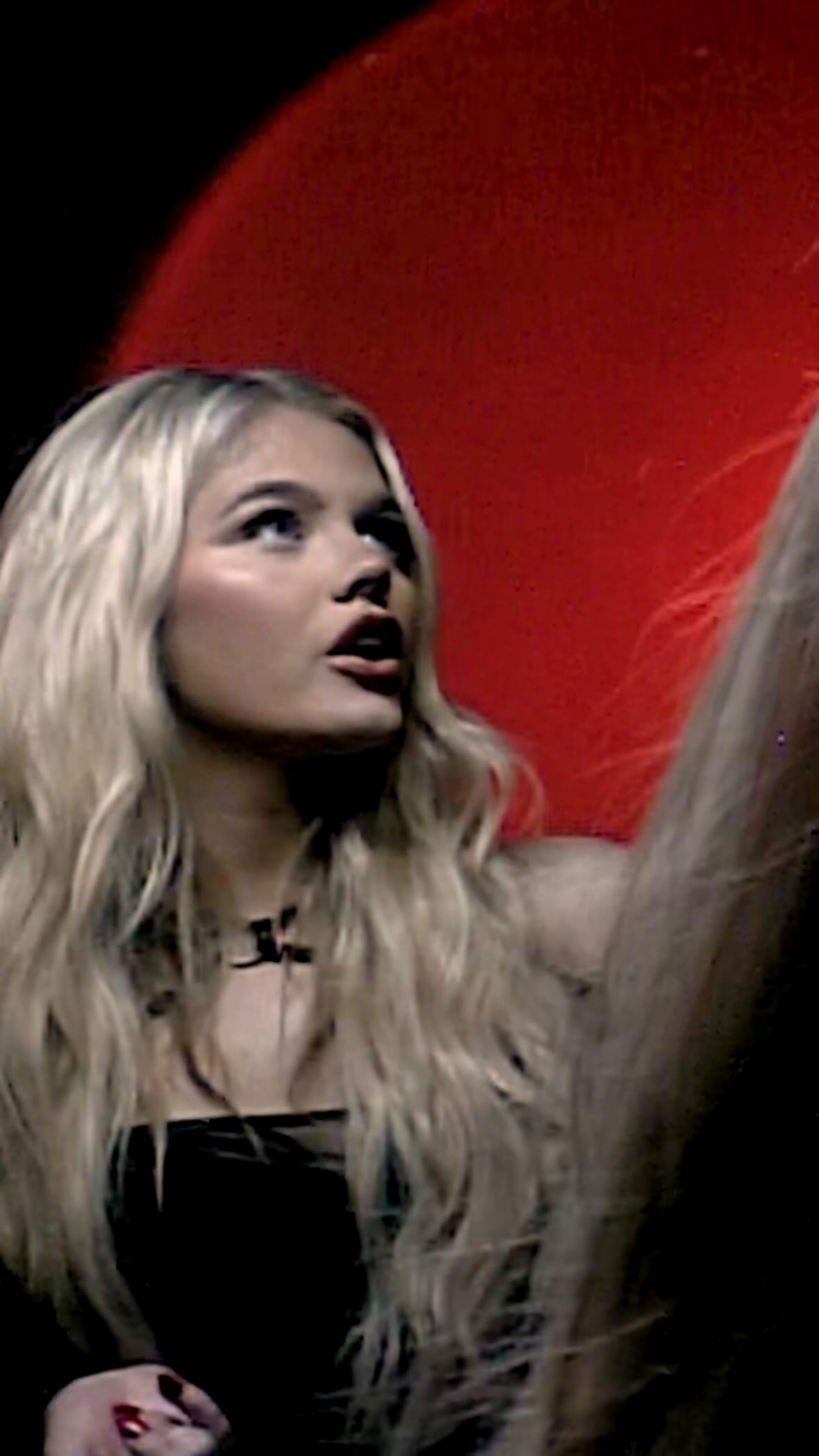 a woman with blonde hair wearing a black dress and necklace looks upward against a red background.