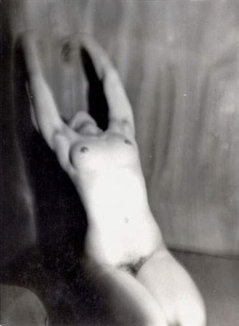 the image is a black and white photograph of a nude female figure. the woman is seated, her arms raised above her head, creating a graceful arc.  the photograph is not sharply focused, having a soft, almost blurry quality that lends an ethereal and artistic feel. the lighting emphasizes the curves of the body, playing with shadows and highlights. the background is out of focus and features a textured surface that suggests fabric or a wall. the overall style suggests fine art photography, possibly from the mid20th century. the pose and the soft focus contribute to a sense of both sensuality and introspection.