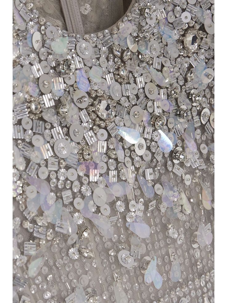 a closeup shot of a sparkly, beaded, and sequined dress. the fabric is a sheer tulle with intricate embroidery of silver and iridescent sequins. the sequins are arranged in a random pattern, creating a shimmering and elegant effect. the dress is perfect for a formal occasion or a special event.