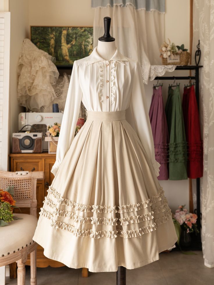 a creamcolored lolita fashion outfit displayed on a mannequin. the outfit consists of a white blouse with delicate ruffled detailing and a long, highwaisted skirt in a lighter cream shade. the skirt features multiple tiers of ruffles along the hem, creating a voluminous and romantic silhouette. the mannequin is positioned in a room with a vintage aesthetic, indicated by the presence of a sewing machine, a clothing rack with various skirts, and a white curtain in the background. the soft, natural lighting enhances the delicate details of the outfit and contributes to the overall elegant atmosphere of the scene.