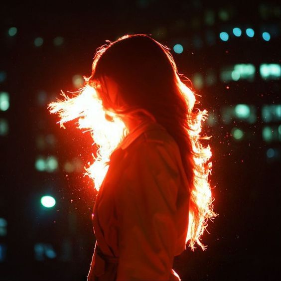 the image shows a woman from her side, standing with her back to the camera. she's surrounded and highlighted by bright orange light that resembles flames, making her hair appear as if it's on fire. the background is a blurry cityscape at night, with specks of light indicating buildings and streets.