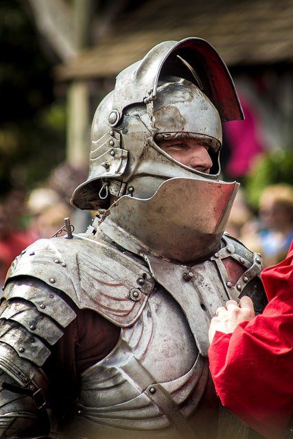 the image captures a closeup view of a knight dressed in full plate armor.  the knight's helmet, with its distinctive raised visor, partially obscures their face, lending an air of mystery and strength. the armor, crafted from gleaming metal, reflects the overcast daylight, highlighting its intricate details and the weight it carries. the red fabric in the foreground likely belongs to another participant, possibly an opponent or an aide. the blurred background suggests a bustling outdoor setting, hinting at a renaissance faire or historical reenactment event.  the overall image evokes a sense of history, power, and the artistry of medieval combat.