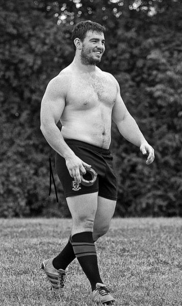 a black and white, fulllength shot of a muscular, shirtless rugby player standing on a grassy field. he's wearing darkcolored shorts with a logo,  striped socks, and cleats. his build is very athletic, and he has a determined expression on his face. the background is blurry, focusing attention on the athlete. the overall mood is one of strength and fitness.