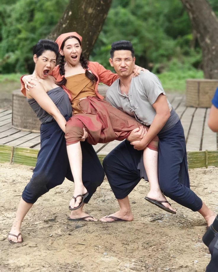 three people are laughing and smiling at the camera while on the beach. two men are carrying the woman, and all are wearing traditional thai clothing. it looks like they are having fun together.