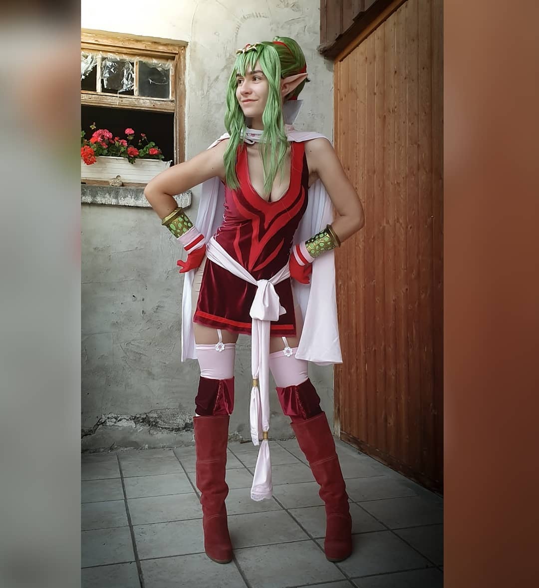 a full shot of a woman cosplaying as lyn from the fire emblem series. she is wearing a red velvet dress with a white cape, pink stockings, and red boots. her hair is styled to match lyn's and she has elf ears. she is posing outdoors, in front of a wooden door and a building.  the background is partially visible, showing some plants and a wall.