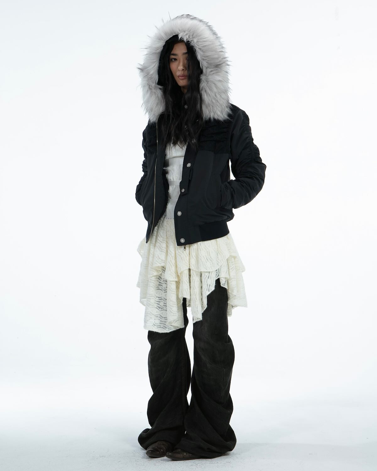 a fullbody shot of a fashion model posing in a black, furhooded jacket, a white ruffled skirt, and black wideleg pants. the model is a woman with dark hair, and she is standing against a plain white background.