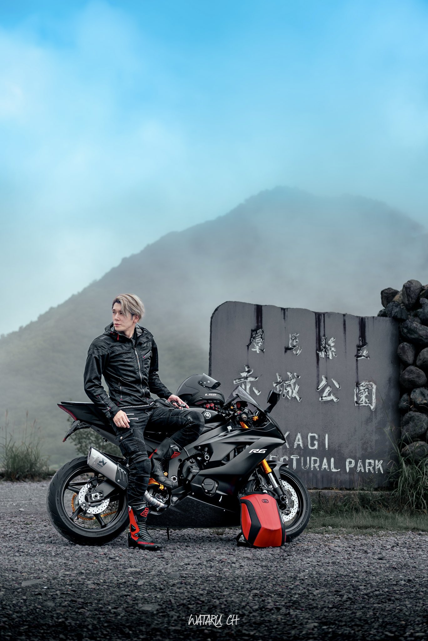 a man wearing a leather jacket and helmet sits on a black sportbike in front of a sign that says "kagi sculptural park" in a scenic mountain landscape. the sky is blue and partly cloudy with fog in the distance. the man is looking off into the distance.