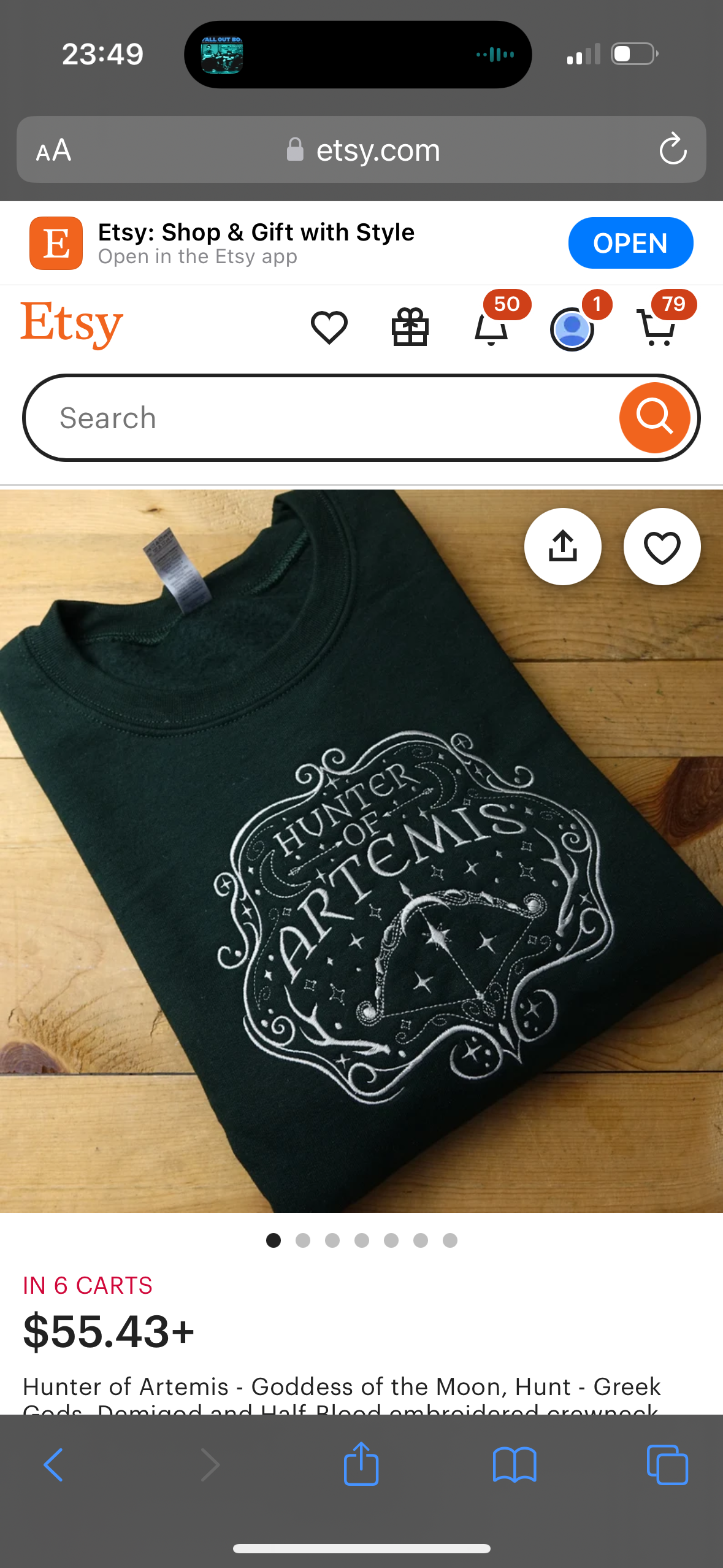 the image is a screenshot of an etsy product page showing a dark green sweatshirt with a white embroidered design of a crescent moon and an arrow, surrounding the words "hunter of artemis" in a script font. the product is titled "hunter of artemis  goddess of the moon, hunt  greek gods demigod and half blood embroidered crewneck" and costs $55.43. the image also shows the etsy logo and the search bar. the page is on a mobile phone. the background is a wooden table.