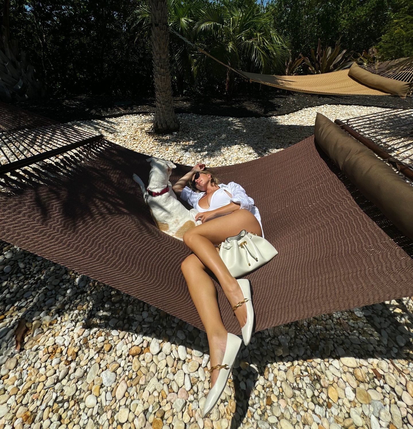 a woman in a white shirt and a white swimsuit is relaxing in a brown hammock with her dog.  she's wearing white shoes and has a white bag next to her.  the hammock is situated on a beach or similar area with small, white pebbles. the scene is sunny and peaceful, suggesting a summer vacation.