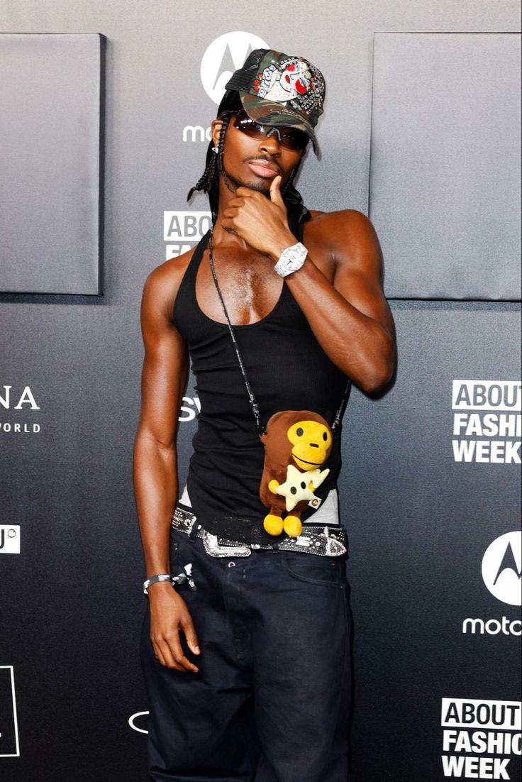 the image shows lil nas x at an about fashion week event. he is dressed in a black tank top, darkwash baggy jeans, a camouflage baseball cap with embellishments, sunglasses, and a bape milo keychain attached to a necklace. he’s accessorized with a watch and a bracelet.  he's posing with his hand on his chin, against a dark background featuring event logos.
