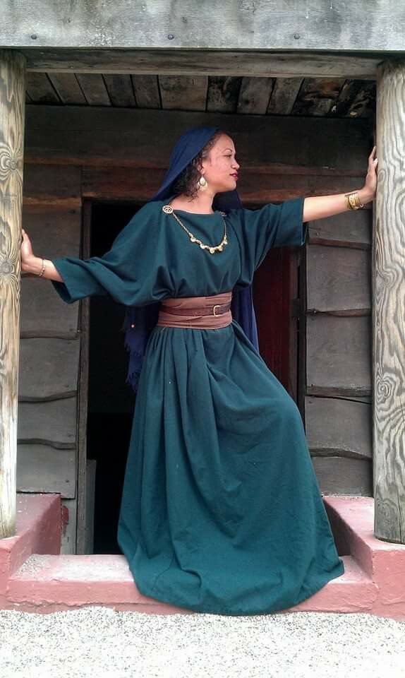 a woman wearing a long, dark green dress evocative of ancient roman attire stands with her hands on wooden posts, looking to the right. the dress is loosefitting with long sleeves and has a brown belt at the waist.  she also wears a dark blue head covering, a necklace, earrings and bracelets. the background is a rustic wooden structure. the overall impression is that of a historical reenactment or photoshoot.