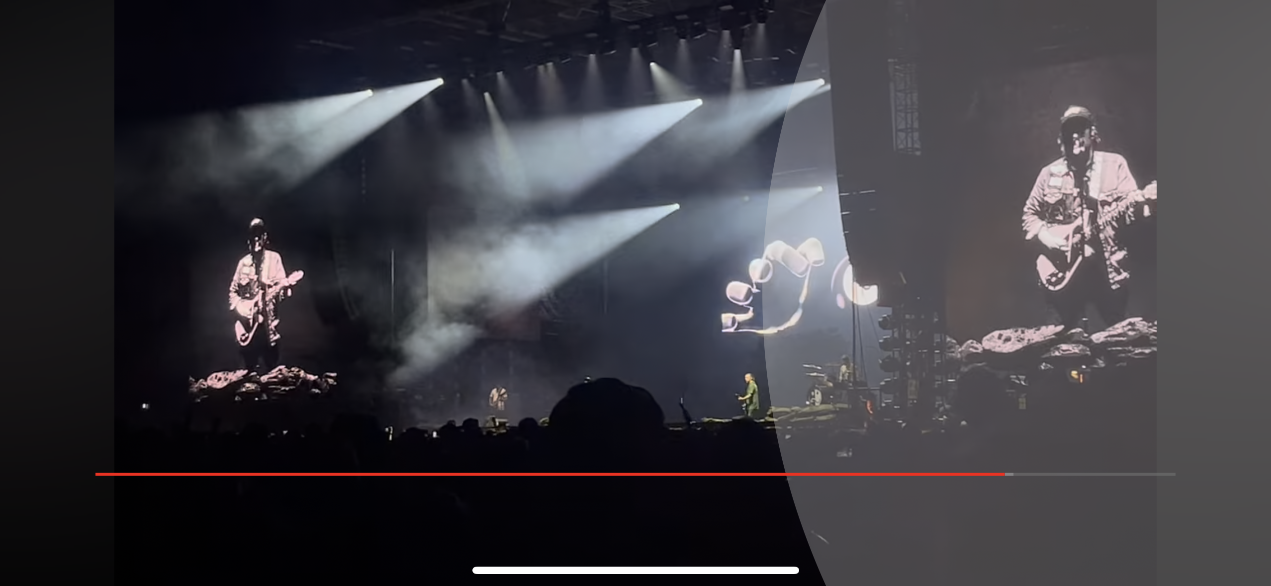a concert stage illuminated with bright spotlights. the audience is in the foreground and the stage is further back. there is a band performing and the lead singer is in the center of the stage, silhouetted in front of a large screen with an abstract, colorful graphic. the lights and shadows of the stage create a dynamic and powerful energy.
