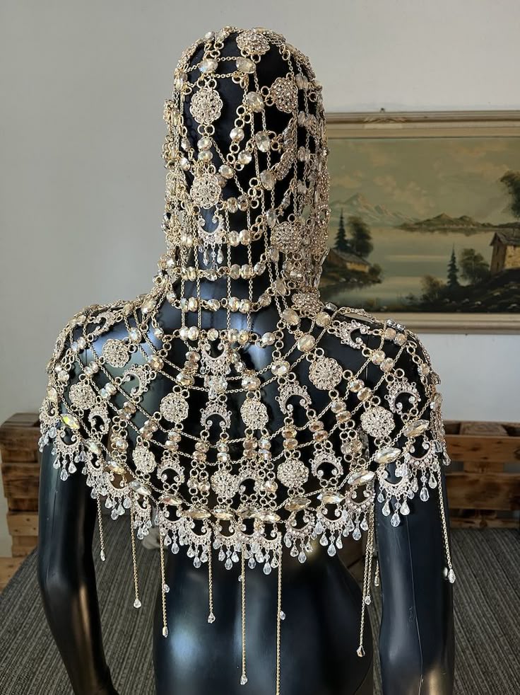 a mannequin is draped with a gold and rhinestone headdress and shoulder piece, creating a veillike effect. the jewelry is intricate with beaded chains, flowershaped elements, and dangling crystals. the background includes a painting and a wooden pallet.