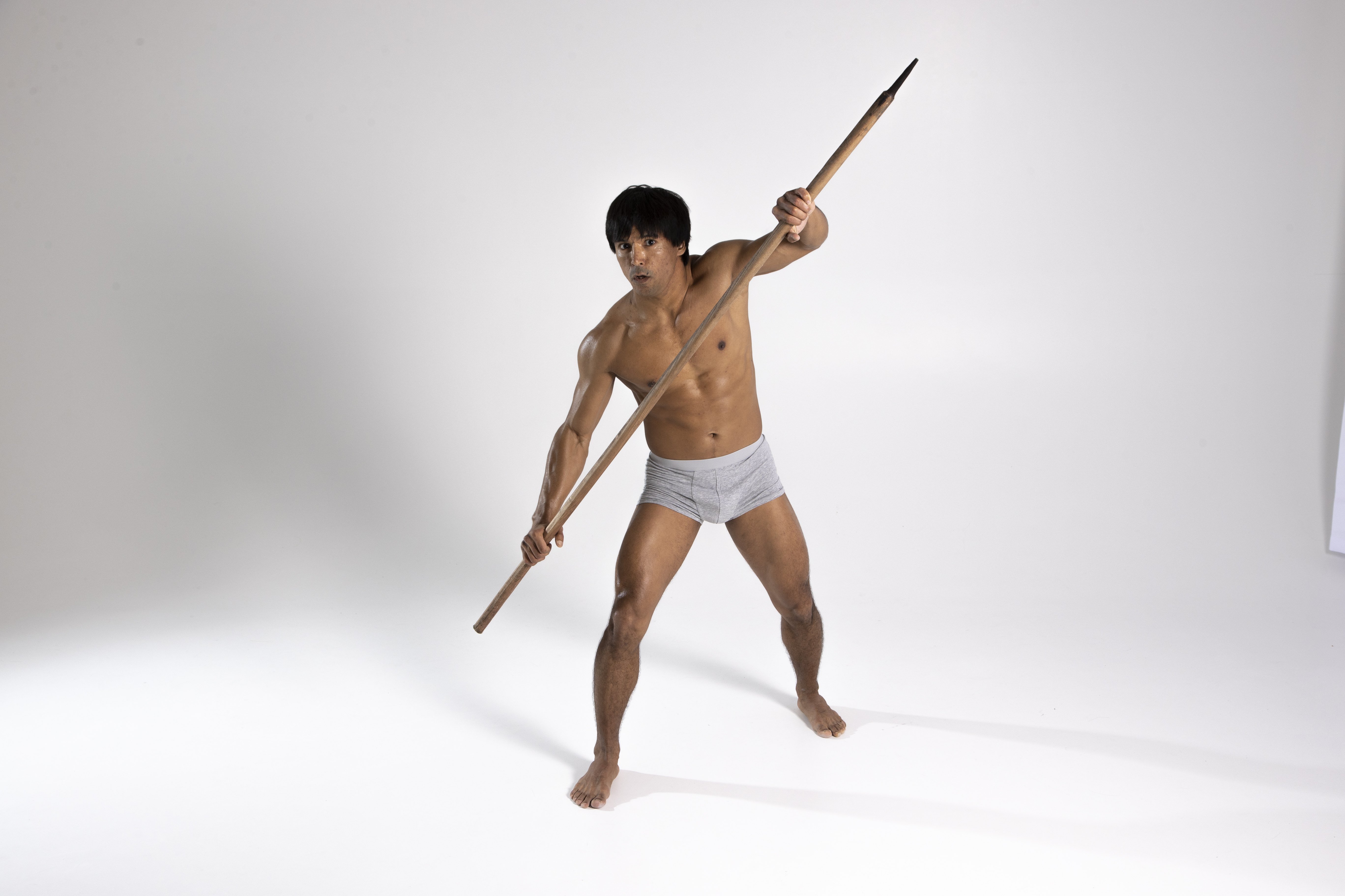 a shirtless muscular man in a loincloth poses with a spear in a studio setting. he is barefoot and has a determined expression.