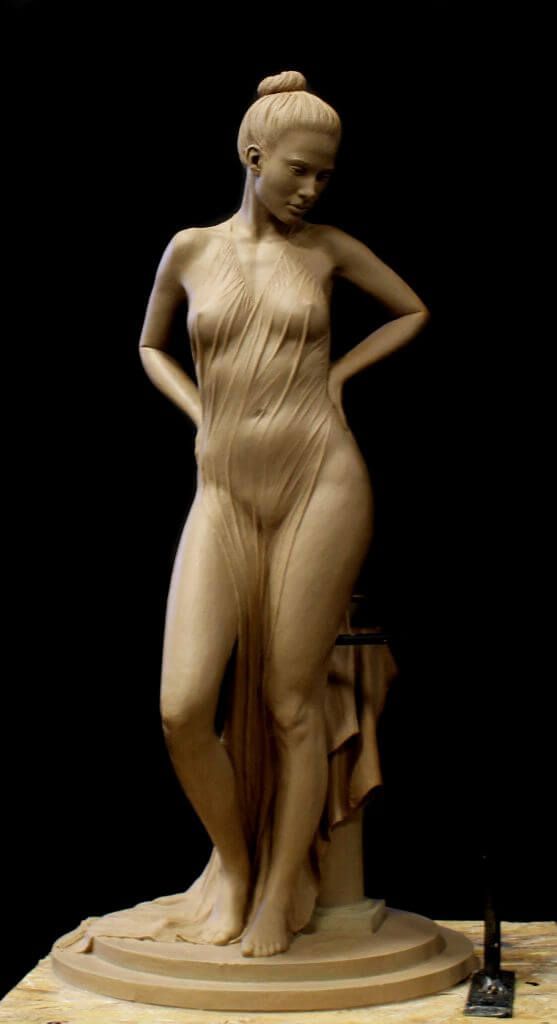 a meticulously sculpted clay figure of a woman stands gracefully on a wooden platform in an art studio. the nude figure is partially draped in a thin, flowing fabric that clings to her body, accentuating her form. the woman has her hair pulled back in a bun and her head tilted slightly downward, her expression contemplative. the lighting is soft and diffused, highlighting the contours of the sculpture and casting subtle shadows. the background is a deep black, focusing the viewer's attention solely on the artwork.