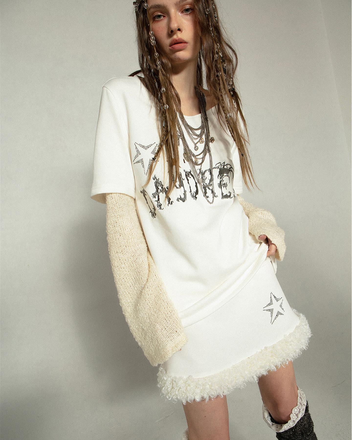woman model wears a stylish offwhite outfit. the outfit consists of an oversized, longsleeved tshirt with a beaded graphic design and a matching mini skirt with a faux fur trim.  the tshirt is layered with a creamcolored knit sweater, and the model is accessorized with multiple necklaces and beaded braids in her hair.  the overall style is bohemian and vintageinspired. the background is a plain lightgrey backdrop that complements the color palette of the outfit.