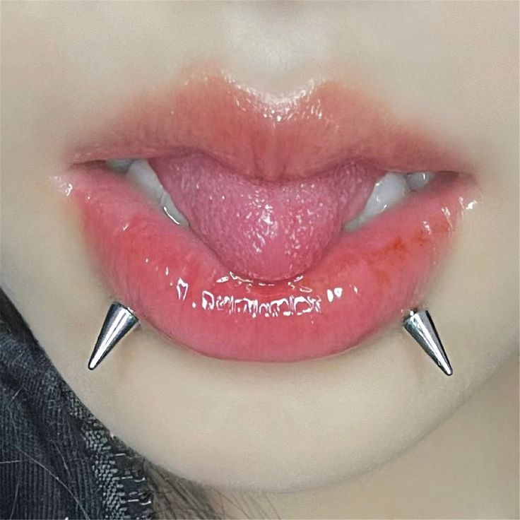 a close up of a person's lower lip with a silver piercing on each side. the lip is glossy and pink, with the tongue slightly sticking out.