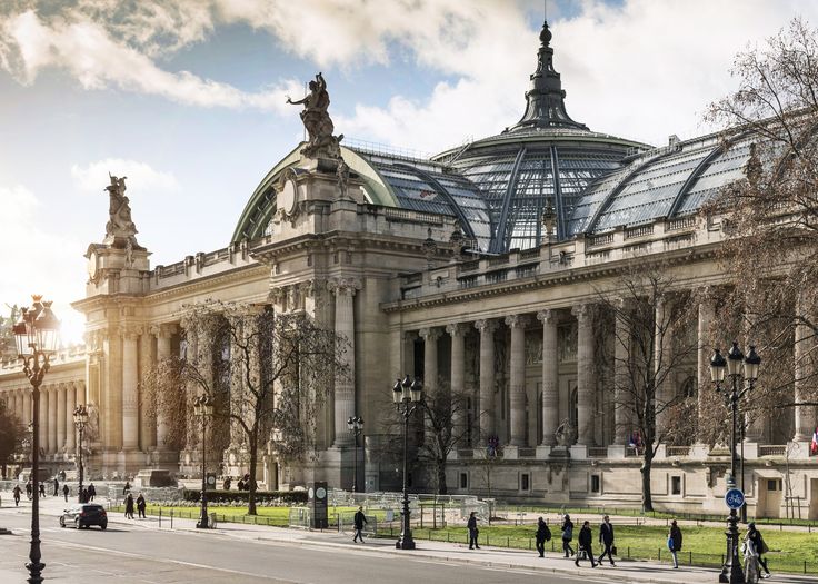 the image showcases the grand palais in paris, france.  the photograph is taken from across the street, offering a full view of the building's impressive facade. the grand, classical architecture is prominent, with numerous columns, statues, and a large glassdomed roof.  the sun is shining, casting light on the building and illuminating the details of its stonework. pedestrians are visible along the street in front, adding a sense of scale and everyday life to the scene. trees line the street, providing some contrast to the imposing building. the overall impression is one of grandeur, historical significance, and the beauty of parisian architecture.