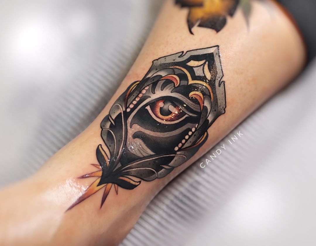a closeup photograph showcasing a freshly done neotraditional tattoo on the back of someone's calf. the tattoo features a stylized eye encased within an ornate, featherlike design. the tattoo is rendered primarily in black and grey, with accents of gold, brown, and white.  the tattoo artist's signature, "candy ink," is faintly visible in the bottom right corner of the image.