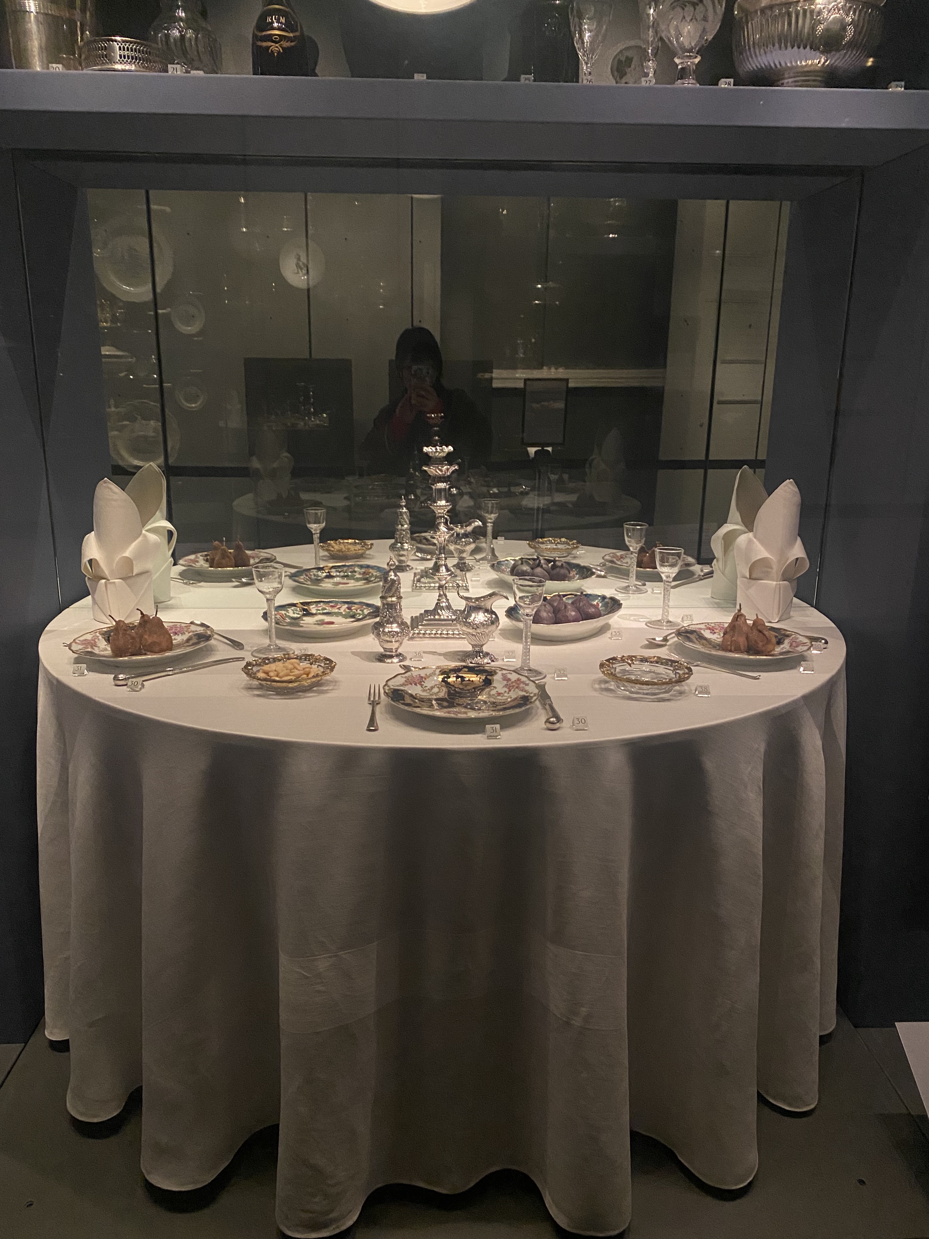 a round table is set for dinner inside a glass display case. the table is covered with a white tablecloth. there are multiple place settings with fine china plates, silverware, glasses, and folded white napkins. food is placed on some of the plates. a silver candelabra sits in the center of the table. the display case is inside a museum or exhibit space. there is a reflection in the glass, possibly of a person taking a photo. a shelf above the display case holds more silverware and dishware.