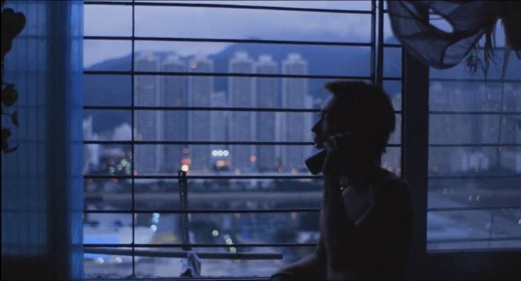 a film still showing a woman silhouetted against a window at night. she appears to be on a phone call, and the view from the window shows a dense hong kong cityscape with many tall buildings. the image is dark, moody, and has a grainy, almost vintage quality, evocative of a film from the 1990s.  the city lights are visible in the distance. the overall atmosphere is melancholy.