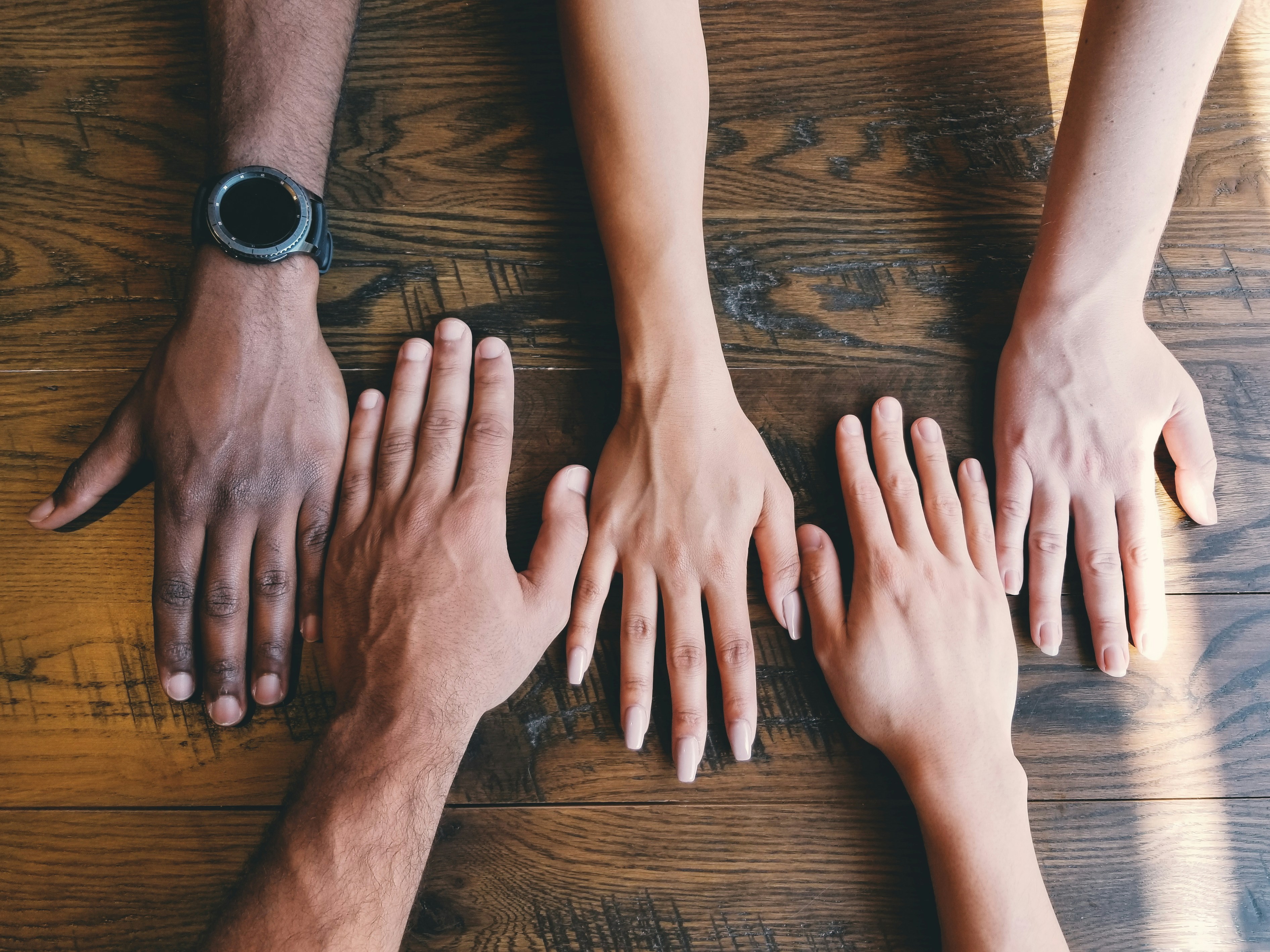 the image showcases the hands of four individuals of different ethnicities lying flat on a wooden table. the perspective is a closeup shot from above, emphasizing the varying skin tones and the texture of the wood. the image is brightly lit, likely by natural light, which creates distinct shadows that accentuate the threedimensionality of the hands.