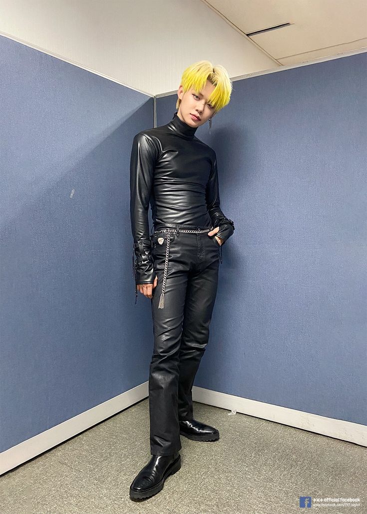 a fulllength shot of a korean man with yellow hair, wearing a black leather turtleneck shirt, black leather pants, and black leather boots, posing in a studio. he has a silver chain accessory on his pants.