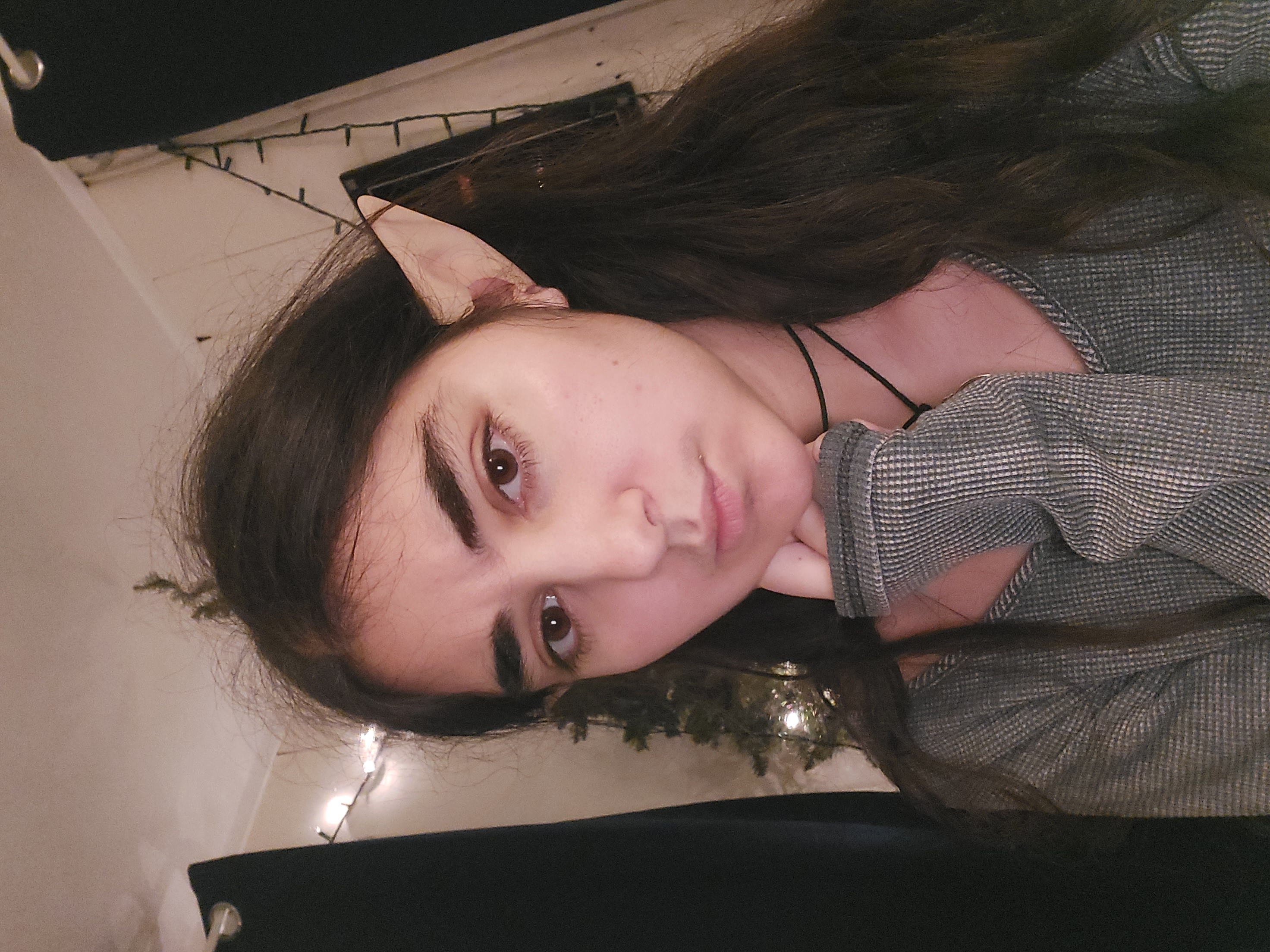 a closeup selfie of a young woman with long, wavy brown hair. she's wearing elf ears, suggesting a cosplay or costume, and a gray thermal sweater.  a simple black choker necklace is also visible.  her expression is serious or pensive. the background is dimly lit, with christmas lights and a partially visible christmas tree. the overall mood is calm and intimate, like a personal snapshot.