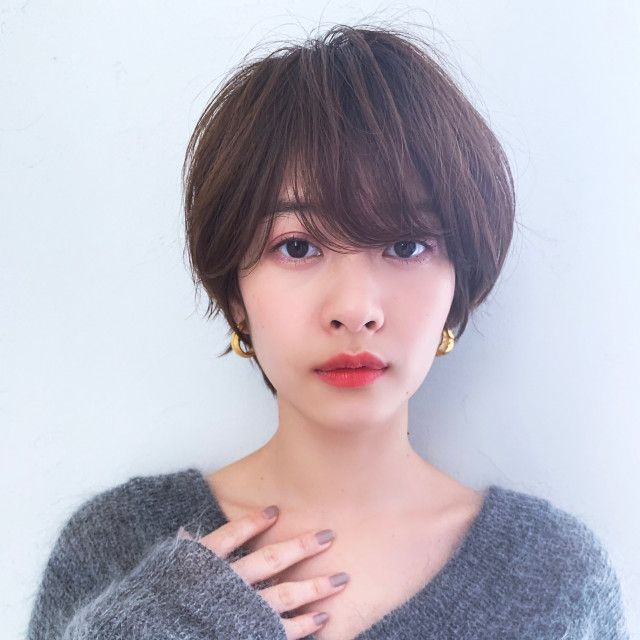 a closeup portrait of a young asian woman with short brown hair and bangs. she is wearing makeup, lipstick, earrings, and a grey sweater. she is looking at the camera with a soft expression.